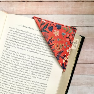 May include: A fabric bookmark with a floral pattern in red, blue, and white, inserted into a book. The bookmark is triangular and has a small tag that reads "APPLES FLUFF". The book is open to a page of text.