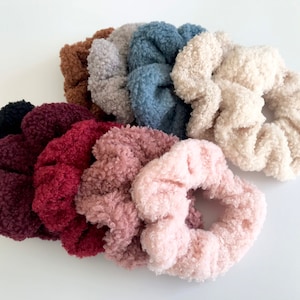May include: A collection of fluffy scrunchies in various colors, including black, burgundy, brown, gray, blue, and pink. The scrunchies have a soft, textured appearance, suggesting a plush material. These hair accessories are suitable for various hairstyles.