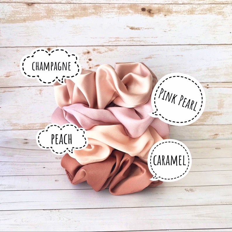 May include: A stack of four satin scrunchies in shades of champagne, pink pearl, peach, and caramel. Each scrunchie is labeled with its color name in a white speech bubble. The scrunchies are arranged on a light wood-grain surface.