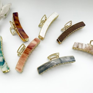 Puede incluir: Una colección de pinzas para el cabello en varios colores y patrones. Las pinzas presentan herrajes metálicos dorados y cuerpos rectangulares curvos en tonos de marrón, blanco, gris y verde. Los patrones incluyen diseños de mármol y carey.
