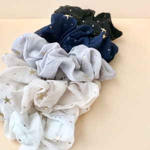 May include: A collection of scrunchies in various colors, including white, gray, navy, and black. Each scrunchie is adorned with gold star accents. The fabric appears to be a lightweight, textured material, suitable for hair accessories.