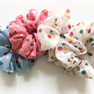 Könnte beinhalten: Vier Stoff-Haargummis in Pastellfarben: Hellblau, Rosa und Weiß. Jedes Haargummi hat ein Muster aus Kirschen und Punkten. Die Haargummis sind aus einem leichten, fließenden Material gefertigt.
