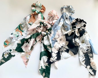 May include: Four floral scrunchies with long, flowing fabric tails. The scrunchies feature a variety of colors, including green, pink, blue, and black, with white floral patterns. The fabric tails add a touch of elegance to these hair accessories.
