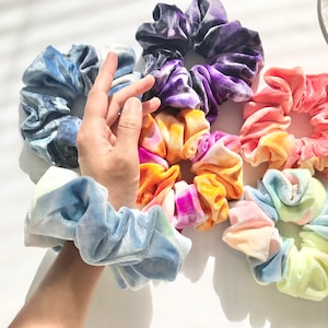 May include: A collection of velvet scrunchies in various colors, including blue, purple, pink, and tie-dye patterns. The scrunchies are arranged on a white surface, showcasing their soft texture and vibrant hues. A hand is visible, wearing one of the scrunchies.