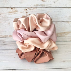 May include: A collection of satin scrunchies in various shades of pink and brown. The scrunchies are stacked together, showcasing the different colors and textures of the fabric. The colors range from light pink to a deep brown.