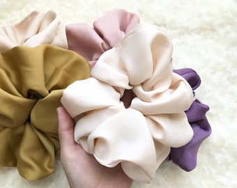 May include: A collection of oversized scrunchies in various colors, including cream, mustard yellow, mauve, and purple. The scrunchies are made of a silky, smooth fabric and are bunched together, showcasing their voluminous design.