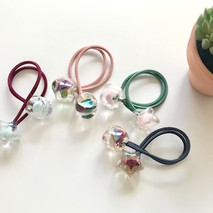 Puede incluir: Colección de coleteros con cuentas transparentes e iridiscentes y detalles en forma de estrella. Las gomas elásticas son de varios colores, como burdeos, rosa, verde y azul marino. Los accesorios están dispuestos sobre una superficie blanca.
