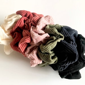 Puede incluir: Una colección de seis coleteros de pana en varios colores. Los coleteros son crema, rosa empolvado, malva, verde oliva, azul marino y negro. La tela tiene una textura acanalada, y los coleteros están agrupados sobre una superficie blanca.