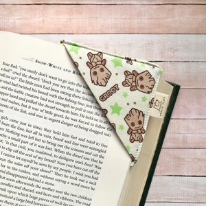 Può includere: Un segnalibro ad angolo bianco e verde con un motivo ripetuto di un personaggio dei cartoni animati e la parola "GROOT". Il segnalibro è inserito in un libro con testo visibile. È attaccata una piccola etichetta con il nome del marchio.
