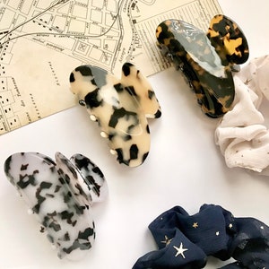 May include: Three hair claw clips in tortoiseshell, cream and black, and black and white patterns. Two scrunchies, one cream with gold stars and one navy with gold stars, are also visible. The items are arranged on a white surface with a vintage map.