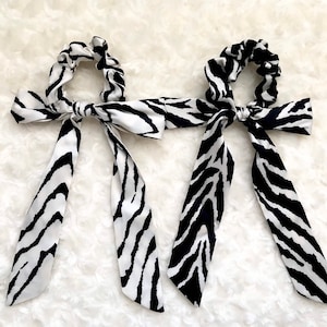 May include: Two scrunchies with long, flowing tails. The scrunchies and tails feature a black and white zebra stripe pattern. The scrunchies are tied with a bow. The background is a white, textured surface.
