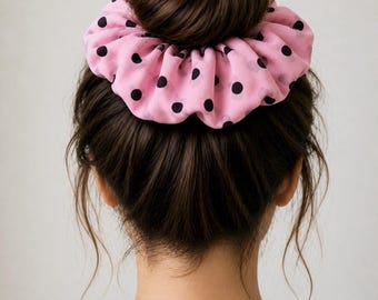 May include: A pink scrunchie with black polka dots sits atop a dark brown bun hairstyle. The scrunchie is made of a soft, gathered fabric. The person is wearing a white top.