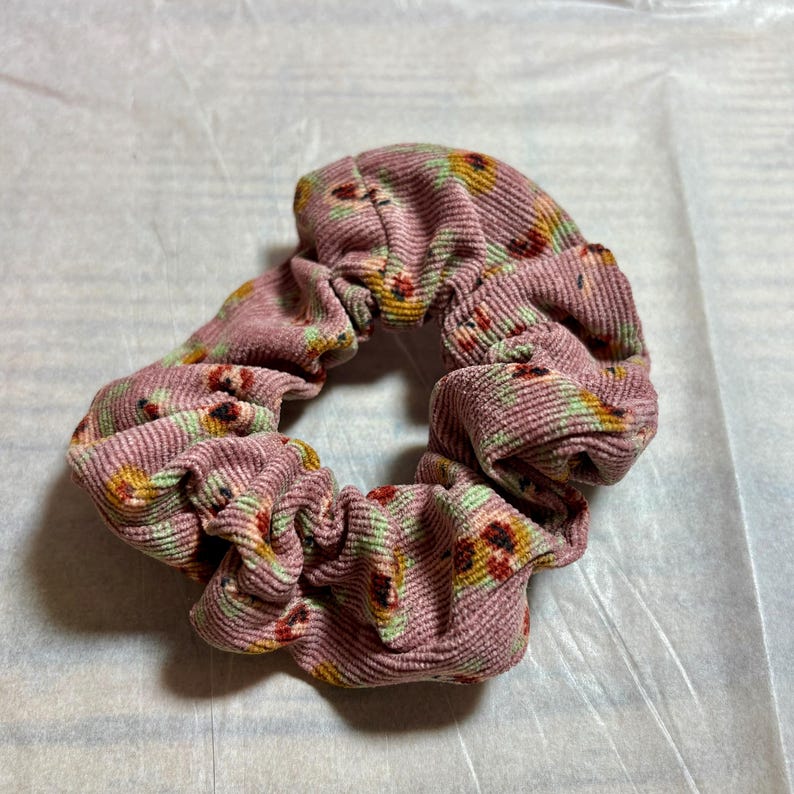 May include: A pink corduroy scrunchie with a floral pattern in yellow, red, and green. The scrunchie is a hair accessory, designed to hold hair in a ponytail or bun.