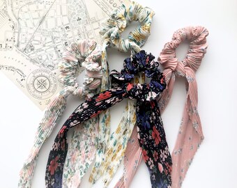 May include: Four pleated fabric scrunchies in various floral patterns and colors, including pink, black, and white. Each scrunchie has a long, matching fabric tail. The scrunchies are arranged on a white surface with a vintage map in the background.