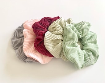 May include: Five pleated scrunchies in a row, each in a different color. The colors are gray, light pink, burgundy, cream, and light green. The scrunchies are made of a soft, textured fabric.