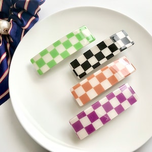 May include: Four rectangular hair clips with a checkered pattern in green, black, peach, and purple. The clips are arranged on a white plate, with a navy blue and white striped bow in the background. These hair accessories are perfect for adding a pop of color.