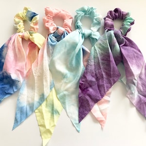 May include: Five colourful scrunchies with long, tie-like fabric tails. Each scrunchie features a different colour scheme, including blue, pink, yellow, and purple, with a tie-dye effect. The fabric tails are attached to the scrunchies.