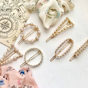 May include: An assortment of gold-toned hair clips, adorned with faux pearls, in various shapes: a circle, triangle, oval, and bar. A map and floral scarf are in the background. The text "Sustainable Acacia Wood Made in Vietnam" is visible.