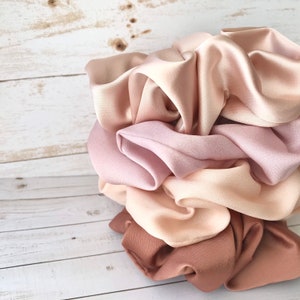 May include: A collection of satin scrunchies in various shades of pink and beige. The scrunchies are bunched together, showcasing the soft, flowing fabric. The colors range from light blush to a deeper mauve, creating a gradient effect.