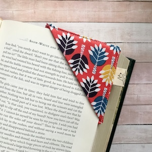 May include: A fabric corner bookmark with a red background and a pattern of blue, white, and yellow leaves. The bookmark is placed on the pages of a book. The bookmark has a small tag that says "APOLLO FLUFF".