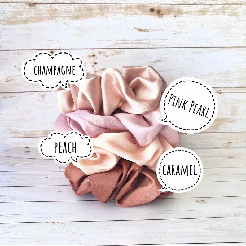 May include: A stack of satin scrunchies in shades of pink, peach, and caramel. The scrunchies are labeled "Champagne", "Peach", "Pink Pearl", and "Caramel". The background is a light wood surface.