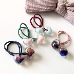 Puede incluir: Colección de gomas para el pelo con forma de corazón en varios colores, incluyendo azul, verde, rosa y morado. Cada goma presenta un charm de corazón transparente e iridiscente unido a una banda elástica a juego. Un lazo de tela malva en el fondo.