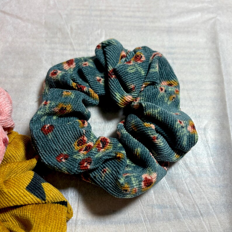 May include: A teal corduroy scrunchie with a floral pattern in red, yellow, and green. The scrunchie is made of a soft, textured fabric and is designed to hold hair in place. Other scrunchies in pink and yellow are partially visible.