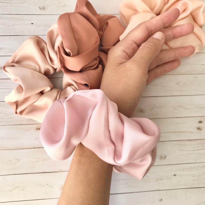 May include: A collection of satin scrunchies in various shades of pink and peach. One scrunchie is worn on a wrist, while others are arranged nearby. The fabric appears soft and has a slight sheen.