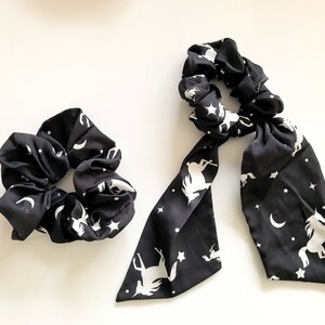 May include: Two black scrunchies with a white celestial pattern of unicorns, stars, and moons. One is a standard scrunchie, and the other has long, flowing fabric extensions.