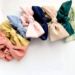 May include: A collection of fabric hair bows in various colors, including pink, yellow, blue, and green. The bows are knotted in the center, creating a decorative accent. The bows are arranged on a white surface.