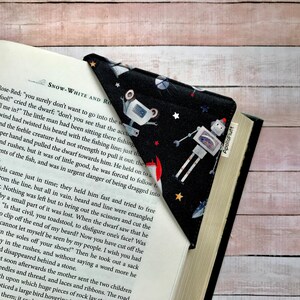 Puede incluir: Un marcapáginas de esquina de tela negra con un diseño espacial de robots, cohetes y estrellas. El marcapáginas está en un libro, con el texto "SNOW-WHITE AND RO" visible.