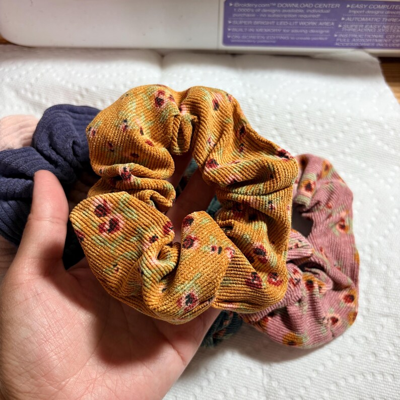 May include: A hand holds a collection of fabric scrunchies. The scrunchies are in various colors, including mustard yellow with a floral pattern, dusty rose with a floral pattern, and navy blue. The scrunchies are made of a corduroy-like material.