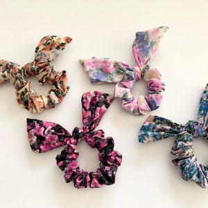 May include: Four floral scrunchies with attached bows are displayed on a white surface. The scrunchies feature various floral patterns in shades of pink, purple, blue, and green. The bows add a decorative touch to the hair accessories.
