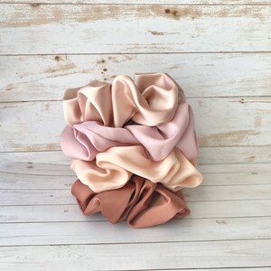 May include: A stack of satin scrunchies in various shades of pink and beige. The scrunchies are stacked on top of each other, showcasing the different colors and textures. The colors range from light pink to a deeper mauve and beige.