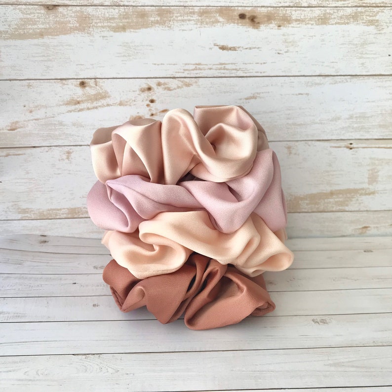 May include: A stack of satin scrunchies in various shades of pink and beige. The scrunchies are stacked on top of each other, showcasing the different colors and textures. The colors range from light pink to a deeper mauve and beige.