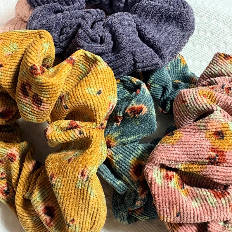 May include: A collection of colorful scrunchies in various shades. The scrunchies are made of corduroy fabric and feature floral patterns in shades of yellow, pink, and teal. Other scrunchies are solid colors, including purple and light pink.