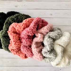 May include: A collection of six fluffy scrunchies in various colors, including black, olive green, coral, pink, light gray, and white. The scrunchies are made of a soft, textured material, likely faux fur or a similar fabric.