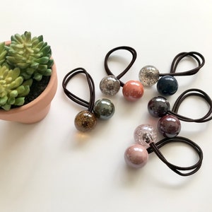 May include: A collection of colorful hair ties with decorative, spherical beads. The beads are various colors, including clear with glitter, brown, pink, and navy. Each hair tie has a dark brown elastic band. A small potted succulent is in the background.