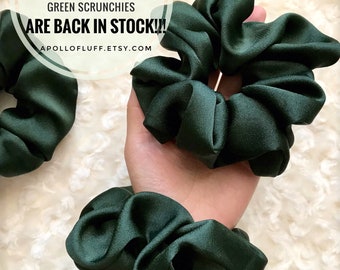 May include: Dark green scrunchies made of a silky fabric. The image shows multiple scrunchies, with one held in a hand. Text in the image reads "GREEN SCRUNCHIES ARE BACK IN STOCK!!" and "APOLLOFLUFF.ETSY.COM".