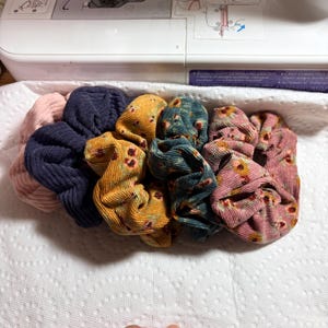 May include: A collection of colorful scrunchies in various fabrics and patterns. The scrunchies include solid colors like navy and pink, and patterned ones with floral designs in shades of yellow, teal, and pink. They are arranged on a white surface.