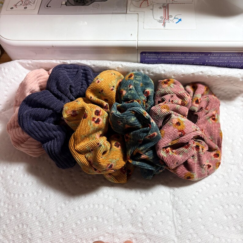 May include: A collection of colorful scrunchies in various fabrics and patterns. The scrunchies include solid colors like navy and pink, and patterned ones with floral designs in shades of yellow, teal, and pink. They are arranged on a white surface.