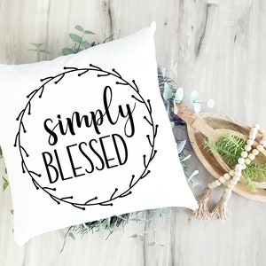 May include: White square pillow with the words "simply BLESSED" in black script inside a black floral wreath. The pillow is surrounded by greenery and wooden decorative elements.