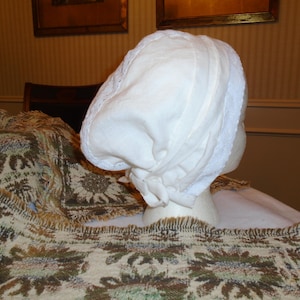May include: A white bonnet with lace trim, likely from the 18th or 19th century, is displayed on a mannequin head. The bonnet is made of a soft, opaque fabric and features vertical seams. The background includes a patterned textile.