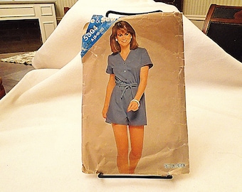 May include: Vintage sewing pattern featuring a woman in a blue romper with short sleeves, a tie waist, and button details. The pattern is labeled "5304 A (8-10-12)" and is displayed on a stand.