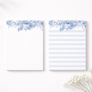 Op de afbeelding: Twee witte notitieblokken met blauwe bloemmotieven. Eén notitieblok is blanco en de andere heeft blauw-witte horizontale strepen. Beide notitieblokken hebben een blauwe bloemenrand aan de bovenkant. Op een witte achtergrond.