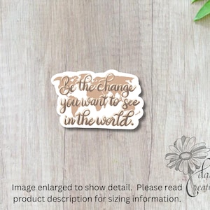 May include: A sticker with the quote "Be the change you want to see in the world" in a handwritten style. The quote is set against a world map design in shades of brown. The sticker is on a light wood grain surface.