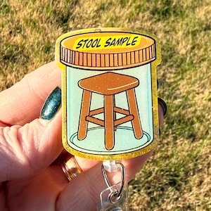 May include: A novelty badge reel featuring a cartoon-style image of a small wooden stool inside a jar. The jar has a brown lid with the words "STOOL SAMPLE" in yellow. The background is a light green color.