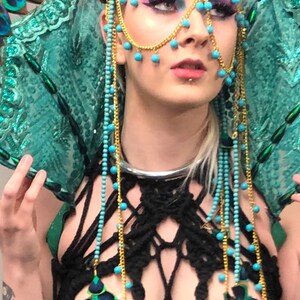 Könnte beinhalten: Eine Frau trägt einen türkis-goldenen Kopfschmuck mit Pfauenfedern. Das Ensemble umfasst ein schwarzes Makramee-Top mit Pfauenfedern-Verzierungen und türkisfarbenem Perlschmuck. Das Make-up beinhaltet lilafarbenen Lidschatten.