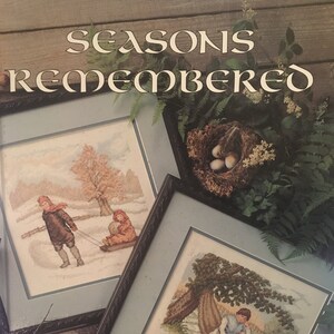 May include: A book cover titled "Seasons Remembered" featuring framed cross-stitch art. The art depicts winter scenes with children sledding and playing in the snow, and a woman reading under a tree. A bird's nest with eggs is also visible.