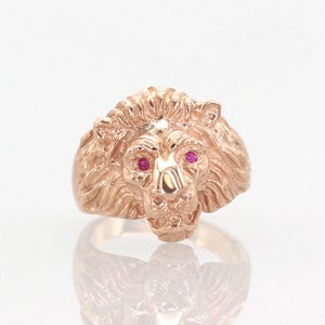 May include: A gold lion head ring with ruby red eyes. The ring is detailed with a textured mane and a snarling expression.